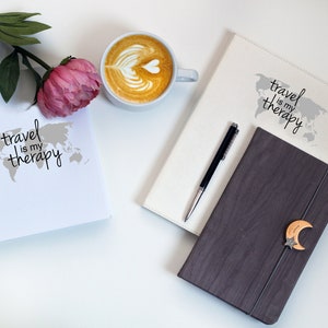 May include: Two white journals with the text "travel is my therapy" and a world map design. A black pen is on top of one journal. A brown journal with a wooden moon and star design is on top of the other journal. A cup of coffee with latte art is in the background.