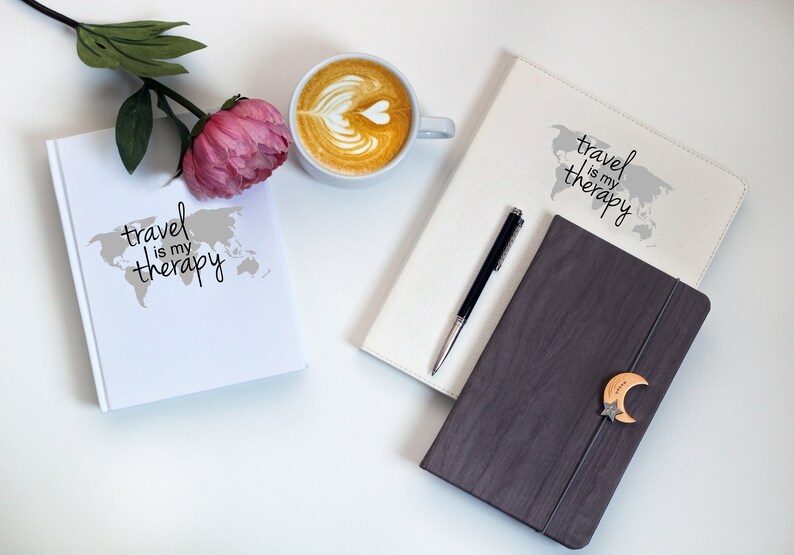 May include: Two white journals with the text "travel is my therapy" and a world map design. A black pen is on top of one journal. A brown journal with a wooden moon and star design is on top of the other journal. A cup of coffee with latte art is in the background.