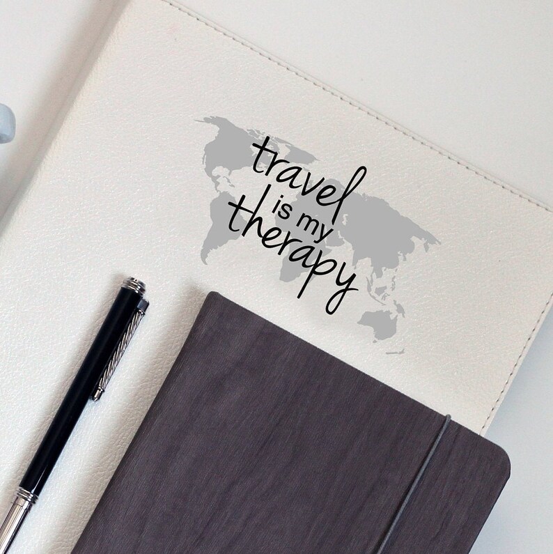 May include: White leather journal with a gray world map and the text "travel is my therapy" in black script. A black pen is lying on the journal. A gray journal with a black elastic band is lying on top of the white journal.