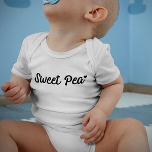 Może przedstawiać: Białe body niemowlęce z napisem "Sweet Pea" czcionką w kolorze czarnym. Body ma kr&oacute;tkie rękawy i okrągły dekolt. Dziecko ma niebieski smoczek.