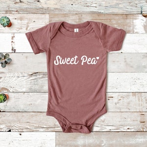 Może przedstawiać: Pudrowy r&oacute;żowy body dla niemowląt z kr&oacute;tkimi rękawami. Na przodzie białą czcionką napisano "Sweet Pea", z małym sercem na końcu. Body wykonane jest z miękkiego materiału.