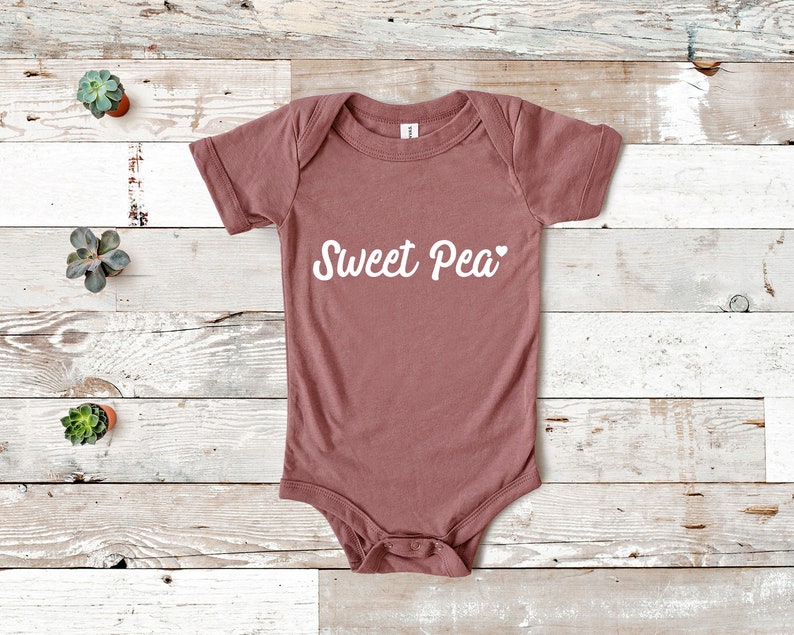 Może przedstawiać: Pudrowy r&oacute;żowy body dla niemowląt z kr&oacute;tkimi rękawami. Na przodzie białą czcionką napisano "Sweet Pea", z małym sercem na końcu. Body wykonane jest z miękkiego materiału.