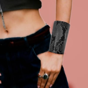 May include: A woman wearing a black sheer top with a black snakeskin bracelet on her right wrist. She is wearing blue jeans and has a silver necklace on.