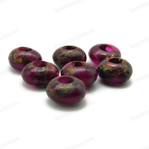 May include: Eight round, dark purple beads with gold flecks and a hole in the centre. The beads are scattered on a white surface.