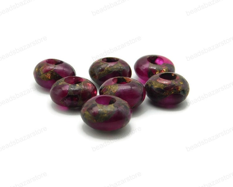 May include: Eight round, dark purple beads with gold flecks and a hole in the centre. The beads are scattered on a white surface.
