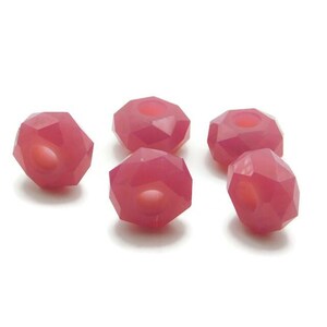Puede incluir: Cinco cuentas de vidrio facetadas de color rosa con un agujero en el centro. Las cuentas están dispuestas en semicírculo sobre un fondo blanco.