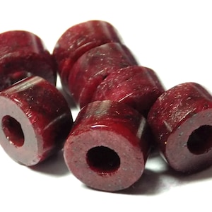 Może przedstawiać: Grupa ośmiu ciemnoczerwonych, okrągłych koralików z otworem w środku. Korale wykonane są z gładkiego, polerowanego materiału.