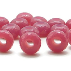 Puede incluir: Un grupo de 15 cuentas de vidrio facetadas de color rosa con un agujero en el centro. Las cuentas están dispuestas en un montón suelto sobre un fondo blanco.