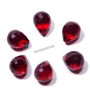 May include: Six faceted, teardrop-shaped beads in a deep red hue are arranged in a circular pattern. The beads have a glossy finish and reflect light, creating a sparkling effect. The text "BEADSBAZARSTORE" is visible.