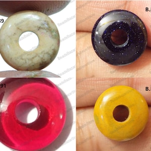 Könnte beinhalten: Vier donut-förmige Perlen in verschiedenen Farben. Eine ist beige mit grünen und braunen Markierungen, eine weitere ist dunkelblau mit Glitzer, eine transparente rote und eine gelb-goldene Farbe. Jede Perle hat ein zentrales Loch.