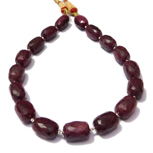 Op de afbeelding: Een streng diepe rode robijn edelsteen kralen, elke kraal is facetgeslepen en ovaalvormig. De kralen zijn geregen op een koord met een gouden en rode kwast aan elk uiteinde.