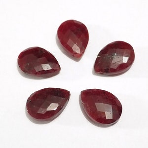May include: Five faceted pear-shaped red gemstones. The gemstones are arranged in a circle on a white surface.