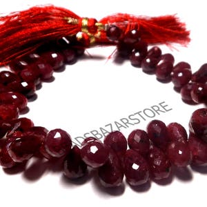 May include: A string of faceted, deep red gemstone beads, possibly ruby, arranged in a curved shape. A red tassel with gold accents is at the top. The beads are on a white background.