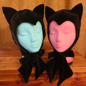 Peut inclure: Deux bonnets de chat en velours noir avec des nœuds en velours noir. Un bonnet est sur une tête de mannequin bleu clair et l'autre sur une tête de mannequin rose.
