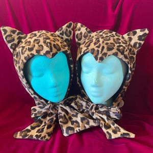 May include: Two leopard print cat ear hoods with matching scarves. The hoods are placed on turquoise mannequin heads against a deep pink background. The hoods have pointed ears and a tie closure.