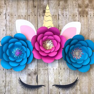 Könnte beinhalten: Ein Einhornkopf aus Papierblumen. Das Einhorn hat eine rosa Blume für das Gesicht, blaue Blumen für die Ohren und ein goldenes spiralförmiges Horn.