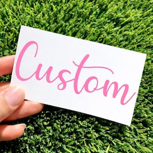 May include: A white rectangular card with the word "Custom" in pink cursive script. The card is held against a backdrop of green artificial turf.