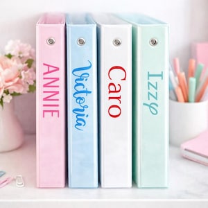 May include: Four pastel-colored binders with names printed on the spines. The binders are pink, light blue, white, and mint green. The names are Annie, Victoria, Caro, and Izzy. The binders are standing upright on a white shelf.