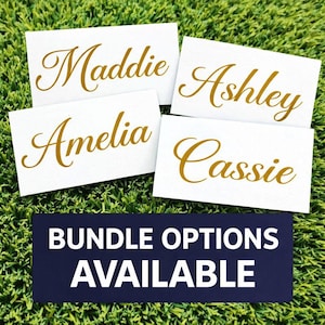 May include: Four white place cards with gold cursive names: Maddie, Ashley, Amelia, and Cassie. A navy blue rectangle below reads "BUNDLE OPTIONS AVAILABLE" in white capital letters. The cards are on a green surface.