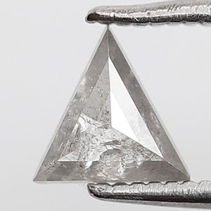 Op de afbeelding: Een driehoekige, heldere, gefacetteerde diamant edelsteen vastgehouden door een pincet. De diamant heeft een ruwe, onbewerkte en ongepolijste uitstraling.