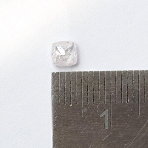 May include: A translucent, cushion-cut gemstone with a faceted surface, positioned next to a metal ruler showing a measurement of approximately 0.25 inches in width. The stone has a pale, cloudy appearance with internal inclusions and sharp edges.