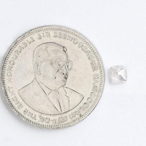 May include: A small, translucent, cushion-cut rough diamond gemstone positioned next to a silver-colored coin. The coin features an embossed portrait of a person wearing glasses and the text THE RIGHT HONOURABLE SIR SEEWOOSAGUR RAMGOOLAM KT.