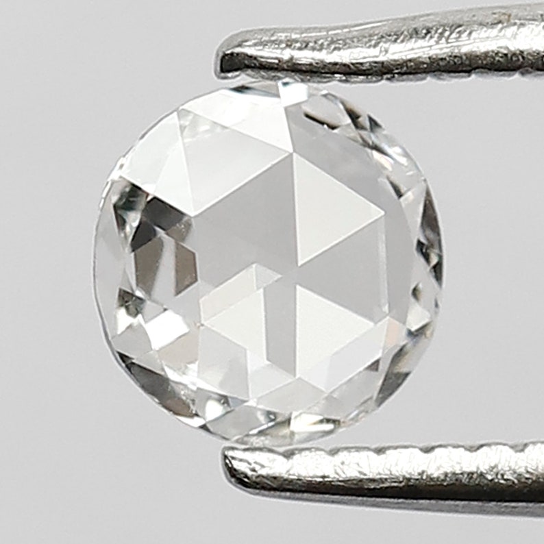 K&ouml;nnte beinhalten: Ein klarer, runder, facettierter Diamant-Edelstein, der von einer Pinzette gehalten wird. Der Diamant hat einen Brillantschliff und ist in einer Zargenfassung gefasst.