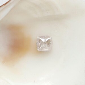 May include: A cushion-cut, translucent white gemstone with a faceted surface, displaying internal inclusions and a clear, crystalline structure. The stone features a square shape with rounded corners and reflects light across its multiple polished facets.