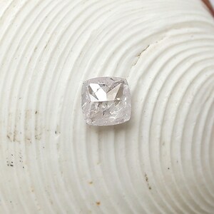 May include: A cushion-cut loose gemstone with a translucent, pale, and icy appearance. The stone features a faceted surface that reflects light, showcasing a square-like shape with rounded corners and a clear, crystalline texture throughout its body.