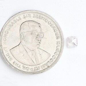 May include: A small, clear, cushion-cut gemstone positioned next to a silver-colored coin. The coin features a portrait of a person wearing glasses and the text THE RIGHT HONOURABLE SIR SEEWOOSAGUR RAMGOOLAM KT.