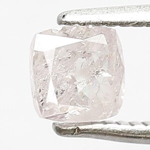 May include: A cushion-cut light pink diamond held by silver-toned metal tweezers. The gemstone features a faceted surface with internal inclusions and a soft, pale rose hue that reflects light across its square-shaped, rounded-corner geometry.