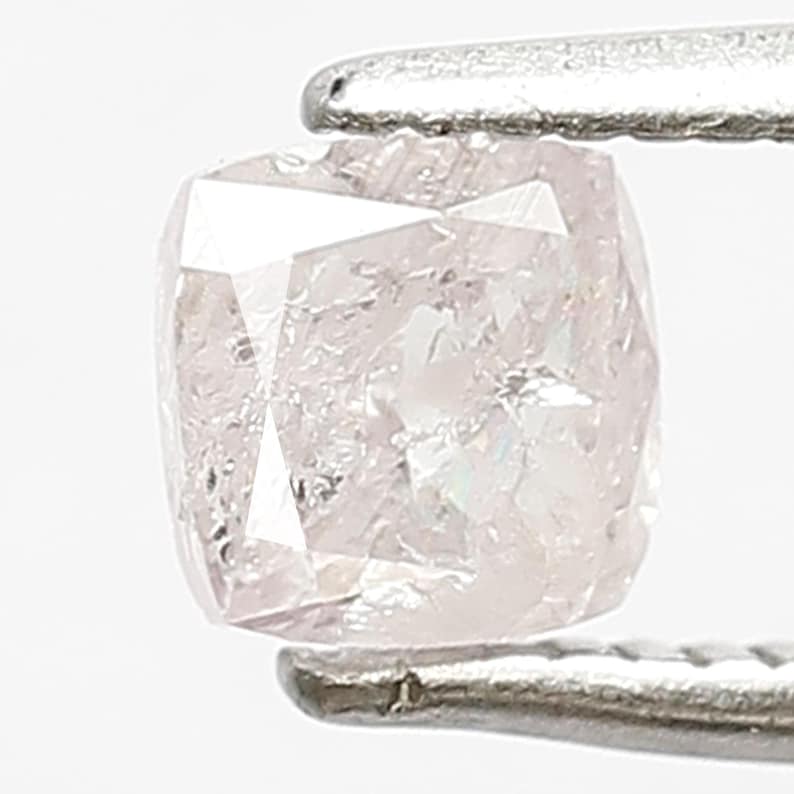 May include: A cushion-cut light pink diamond held by silver-toned metal tweezers. The gemstone features a faceted surface with internal inclusions and a soft, pale rose hue that reflects light across its square-shaped, rounded-corner geometry.