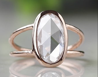 May include: Rose gold ring featuring an oval-cut, faceted, clear gemstone. The gemstone is set within a double-banded rose gold setting. The ring is displayed on a reflective surface, showcasing its design and clarity.