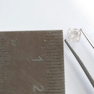 May include: A small, translucent, cushion-cut gemstone with a pale pink hue held by silver-toned metal tweezers. A metal ruler is positioned next to the gemstone, showing it measures approximately 0.125 inches in width and height.