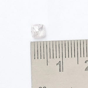 May include: A small, translucent, cushion-cut gemstone with a pale pink hue, positioned next to a metal ruler showing a measurement of approximately 0.25 inches in width, featuring multiple facets that reflect light across its surface.