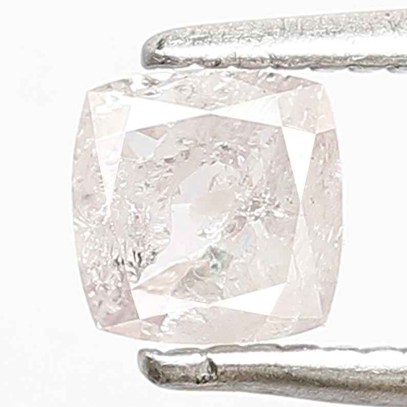 May include: A cushion-cut light pink gemstone held by silver-toned metal tweezers. The stone features a faceted surface with a translucent appearance and a square shape with rounded corners, displaying internal light reflections and clear clarity.
