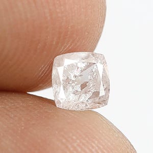 May include: A cushion-cut diamond with a translucent, pale pinkish hue and multifaceted surface. The gemstone features a square shape with rounded corners, displaying internal clarity characteristics and light-reflecting facets throughout its structure.