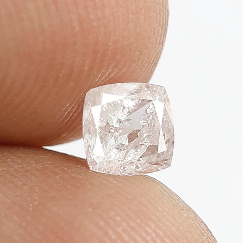 May include: A cushion-cut diamond with a translucent, pale pinkish hue and multifaceted surface. The gemstone features a square shape with rounded corners, displaying internal clarity characteristics and light-reflecting facets throughout its structure.