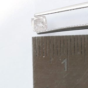 May include: A small, translucent, cushion-cut diamond held by silver-toned metal tweezers, positioned directly above a metal ruler that shows the stone measures approximately 0.2 inches in width and height against a plain white background.