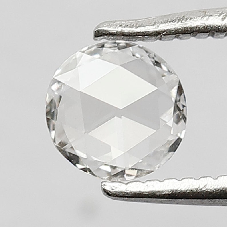 K&ouml;nnte beinhalten: Ein runder, klarer, facettierter Diamant, der von einer Pinzette gehalten wird. Der Diamant ist ein Brillantschliff mit vielen Facetten.