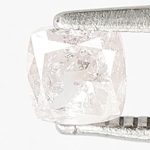 May include: A cushion-cut light pink diamond held by silver-toned metal tweezers. The gemstone features a faceted surface with internal inclusions and a translucent appearance, showcasing a soft blush hue and a brilliant, reflective geometric structure.