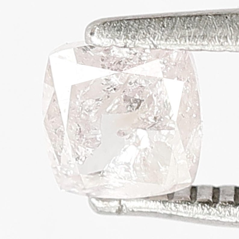 May include: A cushion-cut light pink diamond held by silver-toned metal tweezers. The gemstone features a faceted surface with internal inclusions and a translucent appearance, showcasing a soft blush hue and a brilliant, reflective geometric structure.