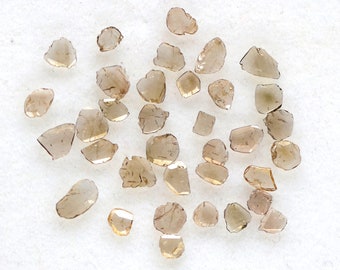 May include: A collection of small, brown, faceted gemstones. The gemstones are scattered on a white surface.