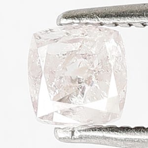 May include: A cushion-cut light pink diamond held by silver-toned metal tweezers. The gemstone features a faceted surface with a translucent appearance, displaying internal reflections and a square shape with rounded corners and clear edges.