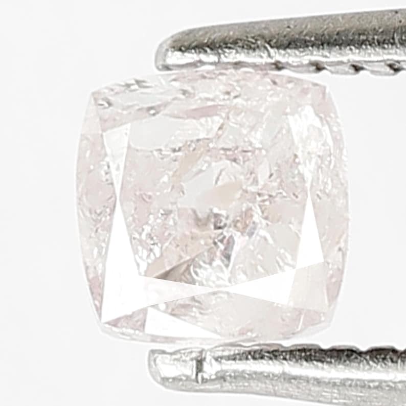 May include: A cushion-cut light pink diamond held by silver-toned metal tweezers. The gemstone features a faceted surface with a translucent appearance, displaying internal reflections and a square shape with rounded corners and clear edges.
