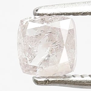 May include: A cushion-cut light pink diamond held by silver-toned metal tweezers. The gemstone features a faceted surface with internal inclusions and a translucent appearance, showcasing a square shape with rounded corners and a soft blush hue.