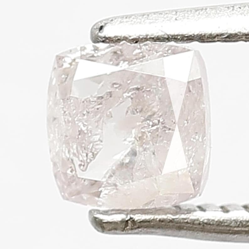 May include: A cushion-cut light pink diamond held by silver-toned metal tweezers. The gemstone features a faceted surface with internal inclusions and a translucent appearance, showcasing a square shape with rounded corners and a soft blush hue.
