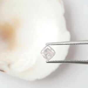 May include: A small, cushion-cut loose diamond with a pale, translucent pinkish hue and visible internal inclusions, held securely by a pair of silver-toned metal tweezers. The gemstone features a faceted surface that reflects light.