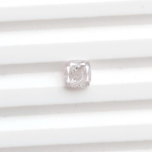 May include: A cushion-cut loose gemstone with a pale, translucent, and slightly pinkish-white hue. The stone features a faceted surface that reflects light, displaying internal inclusions and a clear, crystalline structure throughout the gem.