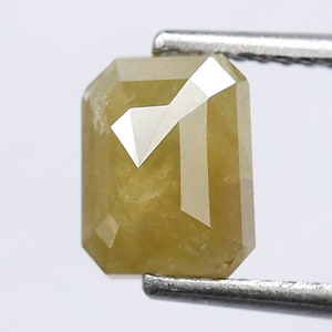 May include: A yellow-green emerald cut diamond with a faceted surface. The diamond is held in place by a pair of tweezers.